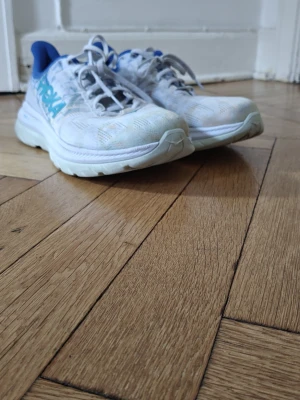 Ljusa Hoka Mach 4 sneakers med blå detaljer - Snygga ljusa sneakers från Hoka One Mach 4  med blå logga och blå insida. Skorna har en sportig design med mesh-ovandel för bra andningsförmåga och tjock, dämpad sula som ger skön komfort. Perfekta för dig som gillar streetstyle och sportig look. Storlek 42 i ny skick 