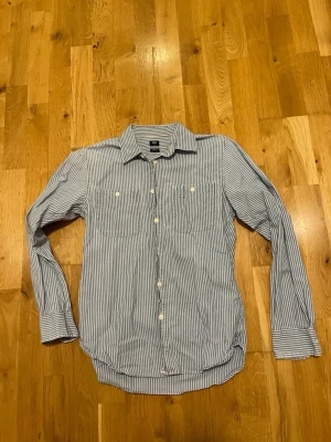 Blåvit randig skjorta från Uniqlo stl XSmall  - Klassisk blåvit randig skjorta från H&M med två bröstfickor och vita knappar. Skjortan har lång ärm, krage och är tillverkad i bomull. Perfekt för en stilren och avslappnad look.