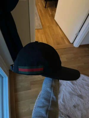 Gucci keps - Säljer en svart gucci keps med justerbart spänne bak. På sidan finns den ikoniska gröna och röda randen. Kepsen har diskret GG-mönster i tyget och är tillverkad i canvasmaterial. Storlek M passar S
