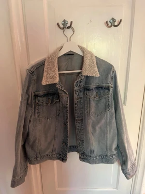 Ljusblå jeansjacka med teddykrage DKNY - Snygg ljusblå jeansjacka från DKNY JEANS med mysig teddykrage. Klassisk modell med två bröstfickor och knappar framtill. Perfekt för dig som vill ha en cool och tidlös look med extra komfort runt halsen. Passar en S/M i storlek. 