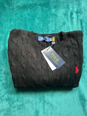 Svart kabelstickad tröja Polo Ralph Lauren - Kabelstickad svart tröja från Polo Ralph Lauren med klassisk röd broderad logga på bröstet. Tröjan har rund halsringning och ribbade muddar. Perfekt för dig som gillar stilrena och tidlösa plagg.