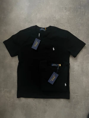 Svart t-shirt från Polo Ralph Lauren - Klassisk svart t-shirt från Polo Ralph Lauren med den ikoniska vita logon broderad på bröstet. T-shirten har rund hals och korta ärmar, tillverkad i mjuk bomull för en skön och stilren look.