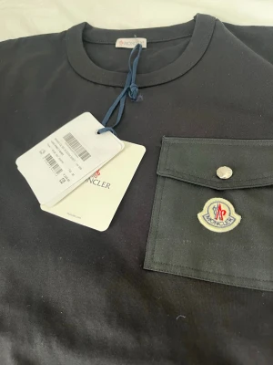 Svart Moncler t-shirt med ficka - Svart t-shirt från Moncler med rund halsringning och en snygg bröstficka med lock och knapp. På fickan sitter Monclers ikoniska logotyp. Tillverkad i mjuk bomull, perfekt för en stilren och trendig look.