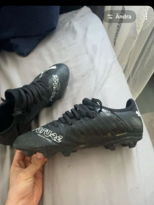 Svarta Puma Future fotbollsskor - Säljer ett par svarta Puma Future fotbollsskor med vit logga och textdetaljer. Skorna har snörning och är tillverkade i syntetmaterial med en modern, sportig design. Perfekta för dig som vill ha stilrena och funktionella skor på planen. Priset kan gå ner om du frågar om det 