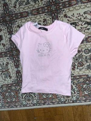Rosa Hello Kitty t-shirt från FB Sister - Söt ljusrosa t-shirt från FB Sister med Hello Kitty-motiv i glittriga stenar på bröstet. T-shirten har rund halsringning och korta ärmar. Perfekt för dig som älskar kawaii och vill sticka ut lite extra.