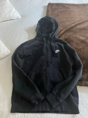 Svart Nike hoodie med dragkedja - Svart hoodie från Nike med dragkedja framtill och vit Nike-logga på bröstet. Utrustad med huva och dragsnören samt två fickor framtill. Tillverkad i mjukt och bekvämt material. Den är i fint skick, hör av er om ni har frågor!