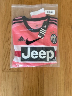 Juventus rosa matchtröja Adidas DYBALA 21 - Juventus matchtröja i rosa med svarta och vita detaljer, tillverkad av Adidas. Tröjan har klubbmärke, Adidas-logga, Nike-logga och Jeep som sponsortryck på bröstet. Rund halsringning och klassiska ränder på axlarna. Perfekt för fotbollsträning eller support. DYBALA 21 på ryggen