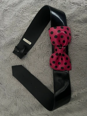 Svart bälte med rosa prickig rosett - Coolt svart bälte med en stor rosa rosett med svarta prickar framtill. Bältet är brett och har en glansig finish som ger en extra touch till outfiten. Perfekt accessoar för att lyfta din look med lite färg och attityd.