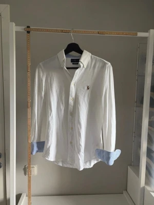 Vit skjorta från Ralph Lauren - Klassisk vit skjorta från Ralph Lauren med broderad logga på bröstet. Långa ärmar med blå- och vitrandiga detaljer på insidan av manschetterna. Skjortan har normal passform och knäppning framtill. Perfekt för en stilren look.
