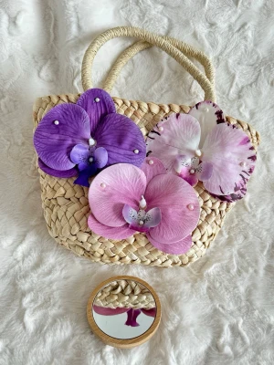 Stråväska med stora blommor - Söt handväska i flätad strå med två handtag och dekorerad med stora konstgjorda orkidéblommor i lila, rosa och vitt. Väskan har en bohemisk och unik look, perfekt för dig som vill sticka ut. Blommorna är prydda med små pärlor för extra detalj. Med väskan följer en mini spegel som jag själv dekorererat med snäckor och pärlor. 