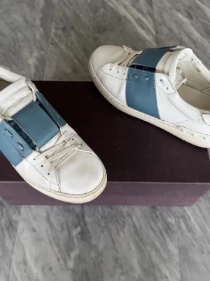 Valentino Garavani vita sneakers - Snygga vita sneakers från Valentino Garavani med bred blå läderdetalj över vristen. Klassisk låg modell med rund tå, vita skosnören och diskreta nitar på hälen. Perfekta för dig som gillar stilrena och exklusiva sneakers.