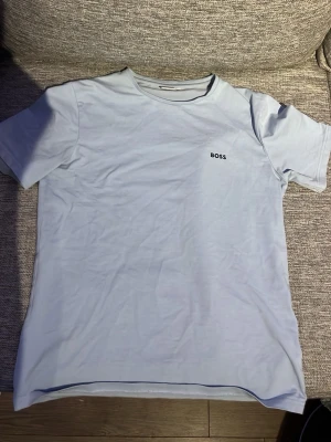 Ljusblå t-shirt från BOSS - Snygg ljusblå t-shirt från BOSS med enkel design och liten logga på bröstet. T-shirten har rund halsringning och korta ärmar, perfekt för en clean och stilren look. Tillverkad i mjuk bomull som känns skön mot huden.