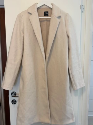 Beige ullkappa från Zara - Stilren beige kappa från Zara med klassisk krage och rak passform. Kappan är något nopprig men det går att få bort. Inga fler bilder skickas då det är packat i flyttlåda.