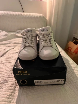 Vita Polo Ralph Lauren sneakers - Snygga vita sneakers från Polo Ralph Lauren med svarta detaljer på hälen och logga på både plös och baksida. Skorna har vita snören med Polo-tryck och är tillverkade i slätt material som ger en clean look.