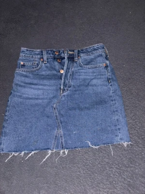 Blå jeanskjol med rå kant - Trendig blå jeanskjol med hög midja, klassiska fem fickor och rå, fransig kant nertill. Kjolen har knappgylf och en lätt A-linjeformad siluett. Perfekt för dig som gillar en avslappnad och cool stil.
