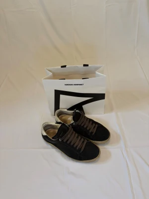 Saint Laurent skor i mocka - Säljer ett par Saint Laurent sneakers i svart mocka med bruna skosnören och vit/beige detalj vid hälen. Skorna är köpta på Nordiska Kompaniet och medföljer NK-påse. Skick: Begagnade men med mycket kvar att ge. Normalt slitage på sula och häl, mockan har naturliga användningsspår. Inlägg och hälparti visar användning. Men sulorna kan enkelt lagas med lite skolim