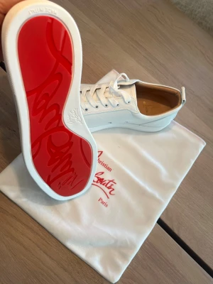 Christian Louboutin vita sneakers - Vita sneakers från Christian Louboutin med ikonisk röd sula och nitar på tån. Skorna har lågt skaft, snörning och är tillverkade i läder. Perfekta för dig som vill sticka ut med en lyxig och edgy stil.