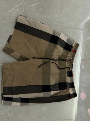 Beige rutiga shorts med snörning - Snygga beige shorts från Burberry med klassiskt rutigt mönster i svart, vitt och rött. Shortsen har elastisk midja med snörning och är tillverkade i ett lätt syntetmaterial. Perfekta för sommaren och ger en trendig look.