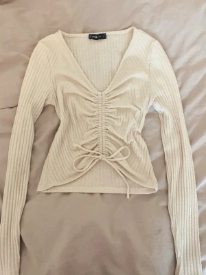 cremevit söt långärmad topp - Trendig beige ribbad topp  med lång ärm, v-ringning och snygg snörning framtill. Omlott-design och croppad passform ger en modern look. Perfekt att styla med jeans eller kjol för en cool outfit.