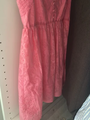 Rosa kort klänning med spetsdetaljer - Söt rosa kortklänning med tunna axelband, spetsdetaljer upptill och knappar framtill. Klänningen har blommigt mönster och dragkedja i ryggen. Perfekt för varma dagar och somriga tillfällen.