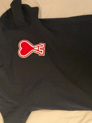 Svart t-shirt från Ami Paris med hjärtdetalj - Svart t-shirt från Ami Paris med stor röd och vit hjärtformad A-logga på bröstet. Klassisk rund hals och korta ärmar. Tillverkad i mjuk bomull som känns skön mot huden. Perfekt för en stilren och trendig look.