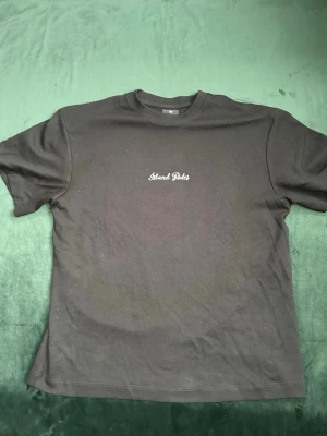 Svart Island Rules t-shirt med tryck - Svart t-shirt från Island Rules med liten vit logga framtill och stort färgglatt tryck med palmer och stadssilhuett på ryggen. Klassisk rund hals och korta ärmar. Perfekt för dig som gillar streetstyle och sköna vibes.