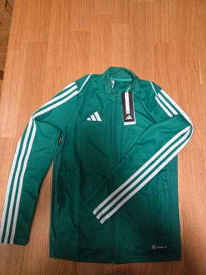 Adidas grön Tiro 23 League unisex träningsjacka, storlek 164 - Grön Adidas träningsjacka med vita ränder längs ärmarna och Adidas-logotypen på bröstet. Jackan har en hel dragkedja framtill och fickor med dragkedja i sidorna. AEROREADY-teknologin är synlig på jackans nederkant.