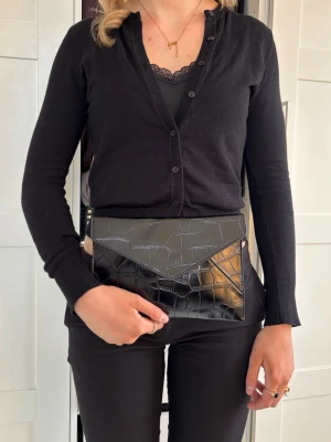 Clutch krokodil mönster  - Med sina mått på 25 x 17 cm är den perfekt stor för det viktigaste – mobil, nycklar och små nödvändigheter – utan att kännas klumpig. Väskan är handgjord i hållbart veganskt läder, vilket ger en mjuk och exklusiv känsla samtidigt som den är ett mer medvetet val.