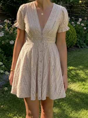 Beige kort klänning från NA-KD - Supersöt beige kort klänning från NA-KD med broderade detaljer och v-ringning. Klänningen har korta puffärmar, knappar framtill. Oanvänd, endast provad!