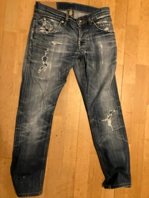 Blå slitna jeans med detaljer - Säljer ett par blå jeans med slitningar och distressed detaljer på både fram- och baksida. Jeansen har klassisk femficksmodell, knappgylf och är tillverkade i denim. Perfekta för en avslappnad och trendig stil.