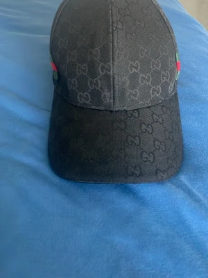 Svart keps från Gucci med monogram - Snygg svart keps från Gucci med klassiskt GG-monogram över hela kepsen. På sidorna finns gröna och röda ränder som ger en lyxig känsla. Kepsen har böjd skärm och är tillverkad i textilmaterial.