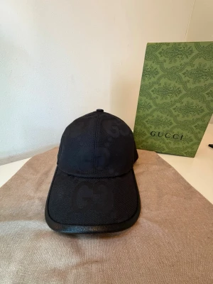 Svart keps från Gucci med GG-mönster - Snygg svart keps från Gucci med det klassiska GG-mönstret ton-i-ton över hela kepsen. Kepsen kommer med box och dustbag. Perfekt till sommaren och matcha din outfit.