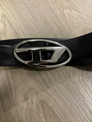 Dieselskärp  - Snyggt svart skärp med bred rem i skinn och en stor, oval silverfärgad buckle med unik design. Perfekt för att ge outfiten en cool touch. Passar till jeans eller byxor för en trendig look.