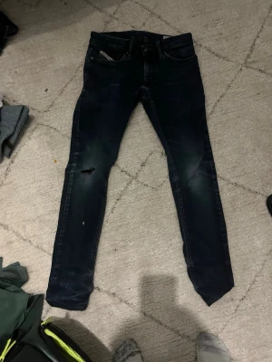 Mörkblå slim fit jeans Shioner - Säljer ett par mörkblå slim fit jeans från Diesel, modell Shioner. Jeansen har stretch, slitningar på ena benet och klassisk femficksdesign. Perfekta för dig som gillar en smal passform och coola detaljer.