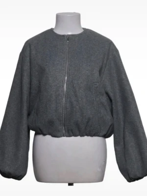 Zara bomberjacka - Snygg grå jacka i oversized modell med rund halsringning och dragkedja framtill. Jackan har ballongärmar och resår i midjan för en avslappnad look. Perfekt för lager-på-lager och streetstyle. Materialet ser ut att vara ullblandning.