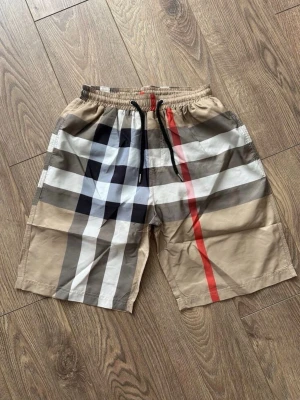 Burrberry Shorts - Snygga beige shorts från Burberry med klassiskt rutigt mönster i svart, vitt och rött. Shortsen har elastisk midja med svart snörning och är tillverkade i ett lätt och bekvämt material. Perfekta för sommardagar när du vill sticka ut lite extra.