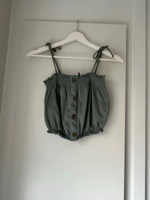 Grön linnetopp med knappar - Snygg grön linnetopp med knytbara axelband och dekorativa knappar framtill. Toppen har resår både upptill och nertill för en puffig och avslappnad look. Perfekt för varma dagar och enkel att styla till jeans eller kjol.
