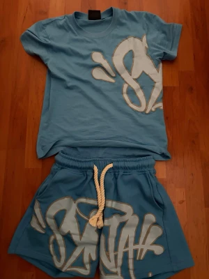 Blått set med graffiti-tryck - Blå Syna World set säljs 💙  Snygg och bekväm Syna World set i ljusblå färg med stora vita detaljer/logga. Setet består av t-shirt och shorts och passar perfekt till sommaren eller vardagsstil.  • Storlek: S • Färg: Ljusblå • Innehåller t-shirt + shorts • Bekvämt material • Trendig streetwear-stil • Fint skick  Hör gärna av dig vid intresse eller frågor.