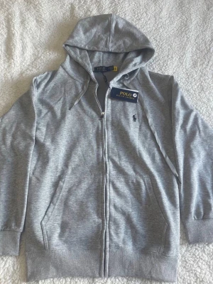 Grå hoodie från Polo Ralph Lauren helt ny - Snygg grå hoodie med dragkedja från Polo Ralph Lauren. Klassisk design med huva, dragsnören och två fickor framtill. Ikoniska Polo-loggan broderad på bröstet. Perfekt för en avslappnad och stilren look.
