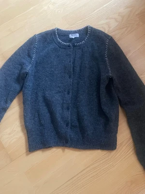 Grå kofta från H&M med vita sömmar - Grå kofta från H&M med rund halsringning och knappar framtill. Koftan har dekorativa vita sömmar längs halsen och axlarna samt ribbade muddar vid ärmslut och nederkant. Perfekt för lager-på-lager och chill stil.