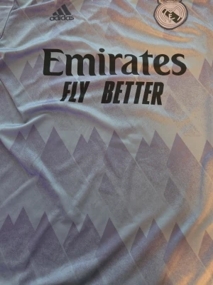 Real Madrid bortatröja Vini Jr 20 - Snygg Real Madrid bortatröja i ljuslila med mönster, tryck av Vini Jr. och nummer 20 på ryggen. Tröjan har klubbmärke och Adidas-logga på bröstet samt sponsortryck framtill. Perfekt för fotbollsfans som vill sticka ut på planen eller läktaren eller på stranden.