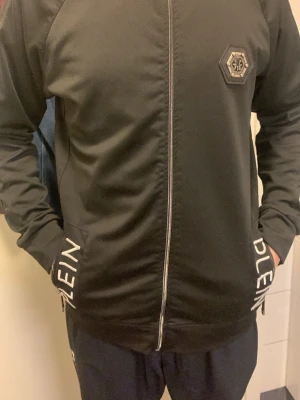 Svart hoodie från Philipp Plein - Snygg svart hoodie från Philipp Plein med dragkedja framtill och vit logotyptext på fickorna. Har ett sexkantigt märke på bröstet och raka ärmar. Tillverkad i ett mjukt material som känns bekvämt och stilrent. Perfekt för dig som gillar streetwear och exklusiva detaljer.