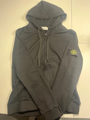 Svart hoodie från Stone Island - Säljer en svart hoodie från Stone Island med dragkedja och klassisk logotyp-patch på vänster ärm. Hoodien har huva med dragsko och ribbade muddar. Perfekt för dig som gillar streetwear och vill ha en stilren look.