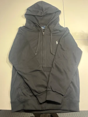 Svart hoodie från Polo Ralph Lauren - Klassisk svart hoodie med dragkedja från Polo Ralph Lauren. Tröjan har huva med dragsko, två fickor framtill och den ikoniska vita loggan broderad på bröstet. Tillverkad i mjuk bomullsblandning för skön komfort och stilren look.