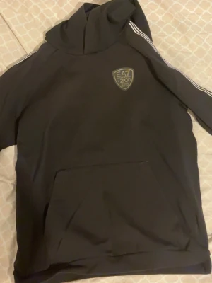 Svart EA7 Emporio Armani hoodie - Svart hoodie från EA7 Emporio Armani med guldiga loggor på bröstet och ryggen. Klassisk huva, känguruficka och vita ränder längs ärmarna. Perfekt för en sportig och trendig look. Materialet känns mjukt och bekvämt. Loggan på bröstet är lite trasig men syns ej när man har på den