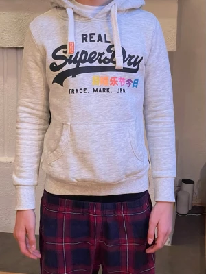Ljusgrå hoodie från Superdry - Ljusgrå hoodie från Superdry med stor logga och japansk text framtill i olika färger. Perfekt för en avslappnad och trendig stil. Jag är ca 172 här och den är lite för liten för min smak. Så jag tror att den sitter som bäst på de flesta mellan 160-170. Det är bara att skriva om ni har frågor! (Priset kan diskuteras)