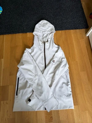 Vit vindjacka med huva från Moncler - Snygg vit vindjacka från Moncler med hel dragkedja, huva och svarta detaljer vid fickor och ärmslut. Jackan har en klassisk Moncler-logga på bröstet och justerbara muddar. Perfekt för blåsiga dagar och enkel att matcha till olika outfits.
