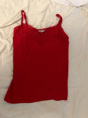 Röd linnetopp med spetsdetalj - Snygg röd linnetopp med tunna axelband och v-ringad spetskant upptill. Perfekt för att lyfta vilken outfit som helst med en pop av färg och romantisk känsla. Toppens material känns mjukt och följsamt mot huden.
