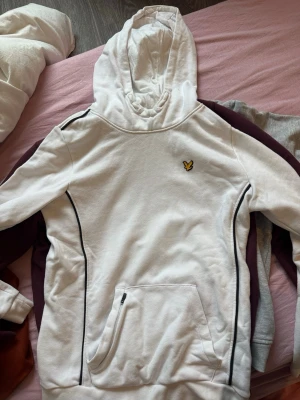 Vit hoodie från Lyle & Scott - Snygg vit hoodie från Lyle & Scott med svart piping längs sidorna och klassisk gul logga på bröstet. Hoodien har en stor huva och en praktisk magficka med dragkedja. Perfekt för en chill och sportig look.