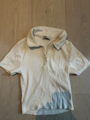 Vit ribbad polotopp från Fila - Klassisk vit ribbad polotopp från Fila med korta ärmar och knappar framtill. Tidlös design med mjukt material och diskret logga i nacken. Perfekt till jeans eller kjol för en sportig look.
